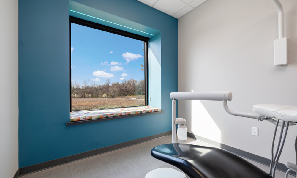Modern dental treatment room with blue accent wall, large window overlooking nature, and dental chair with overhead light