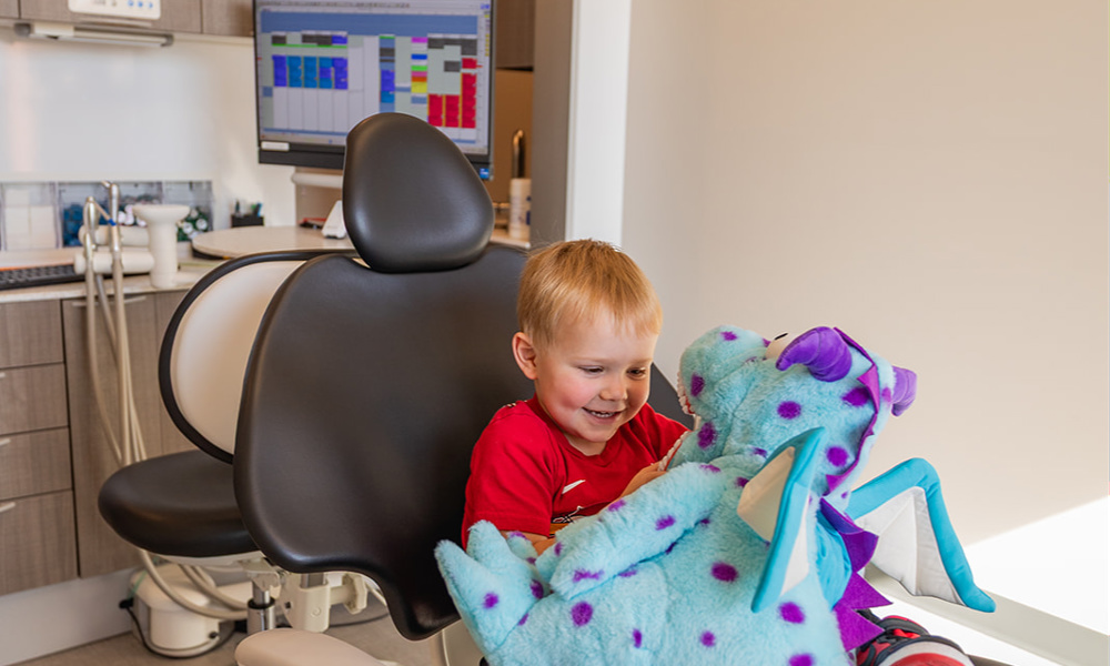 Smiling young boy sitting in dental chair playing with a blue stuffed animal dragon toy